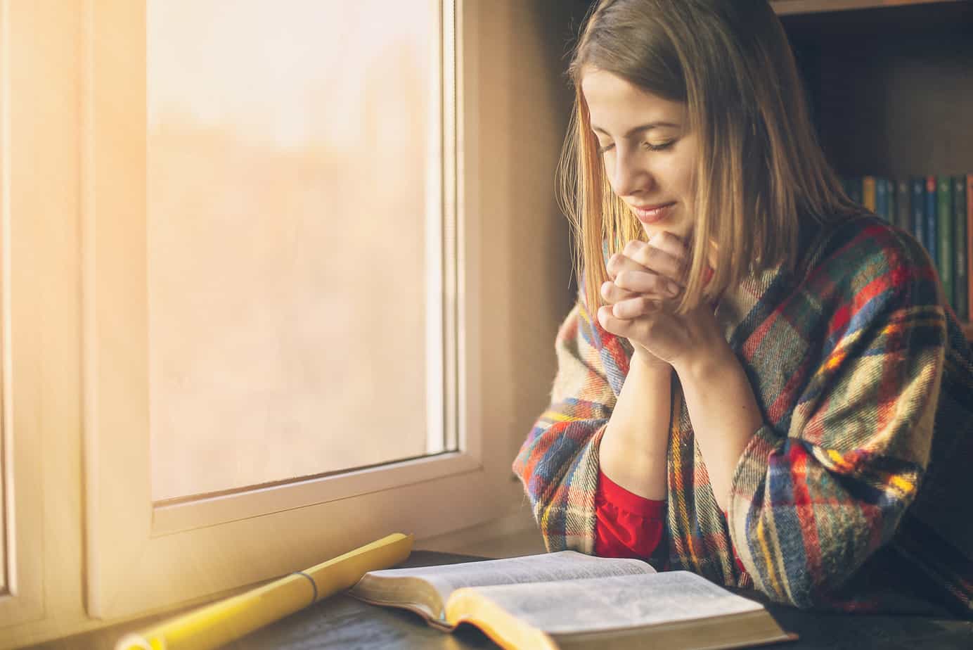prayers for husband - young woman prayers near window with golden light streaming through, open bible in front of her