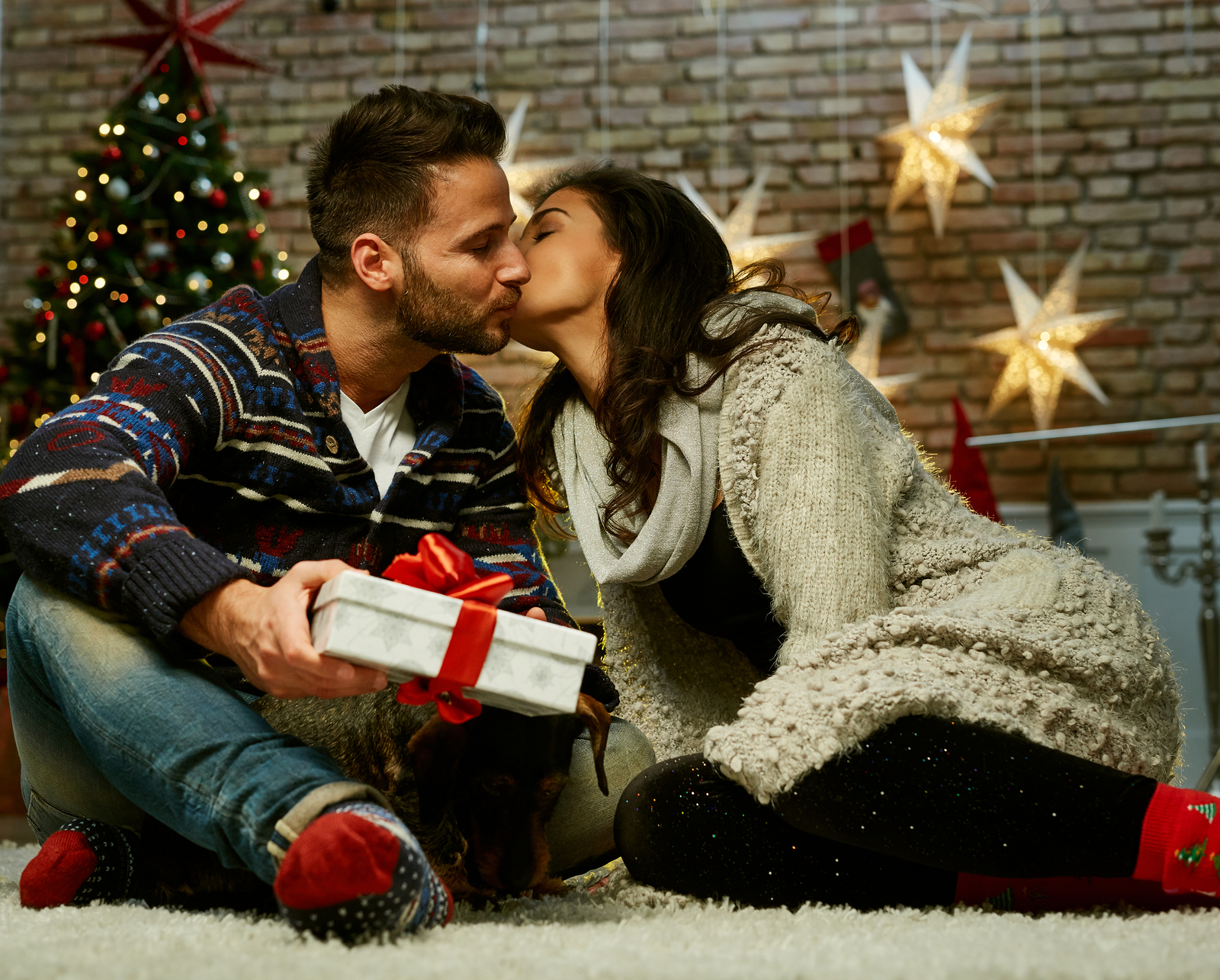 stocking stuffers for husband - Christmas couple at home in Winter. Happy young couple sitting on floor at home with gift box in Christmas time. Christmas tree and fireplace in background.