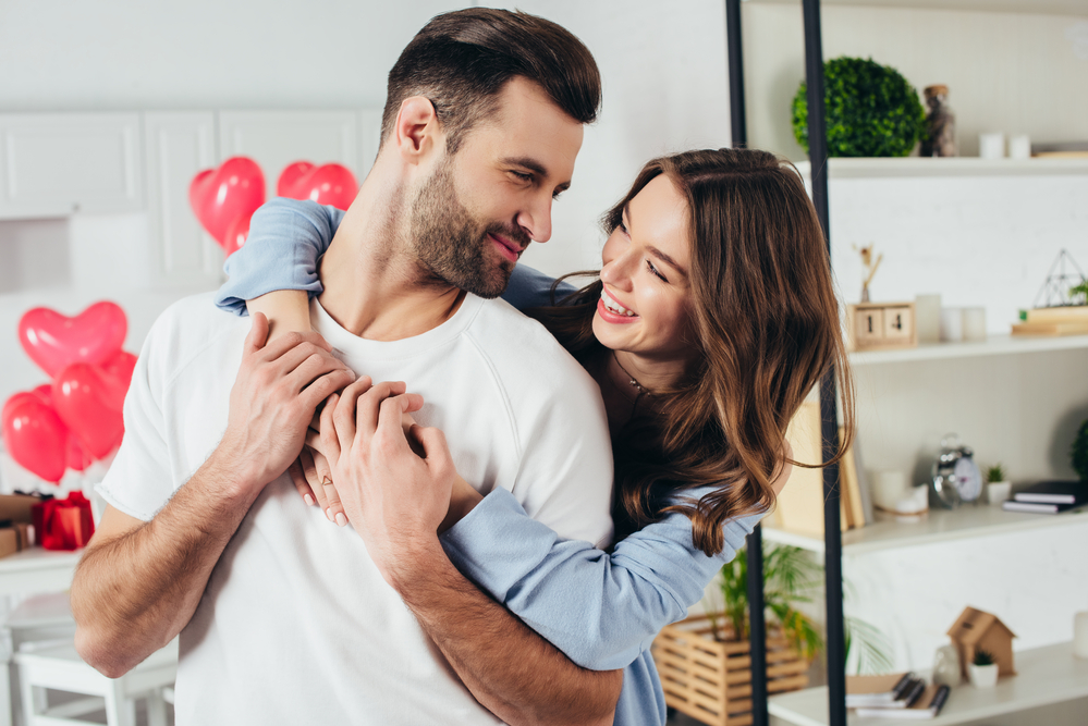 best date night subscription boxes - photo of white couple standing in house smiling, woman hugging husband from behind