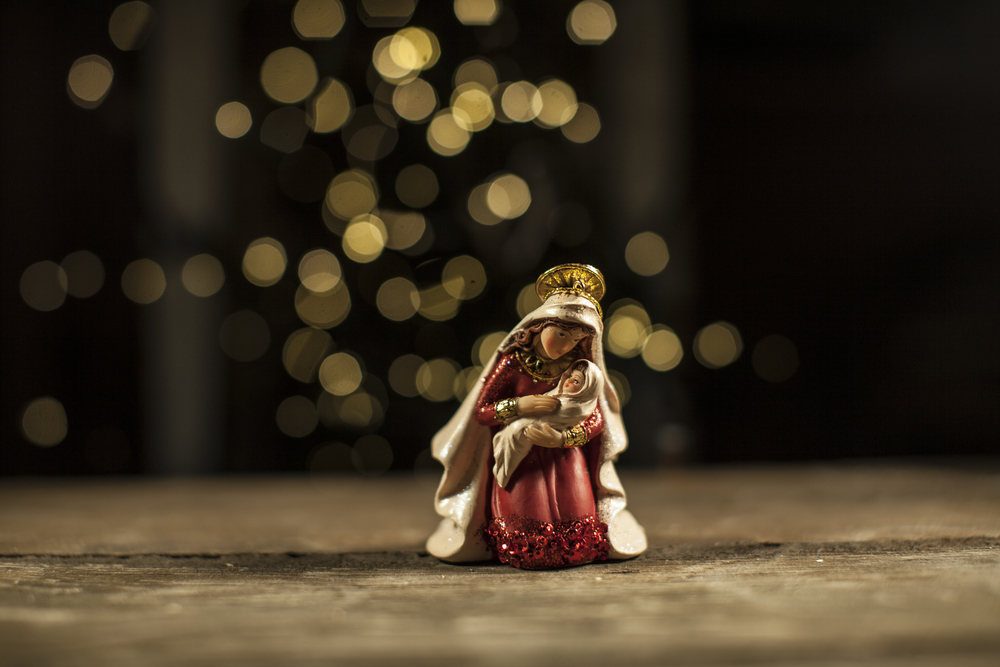 a small statue of mary cradling jesus sits on a table with blurry white christmas lights in the background